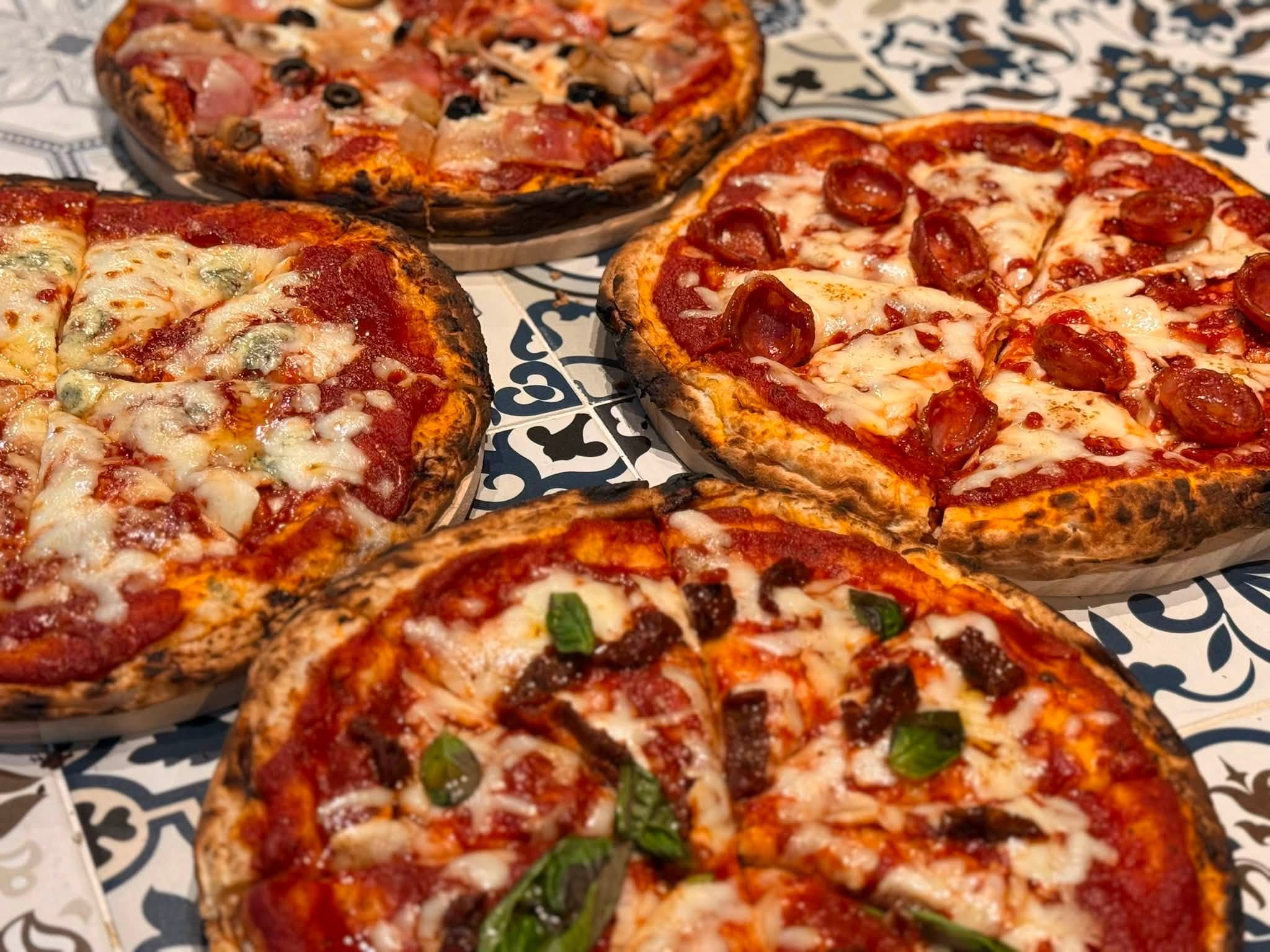 Four wood-fired pizzas with blistered crusts on patterned tiles